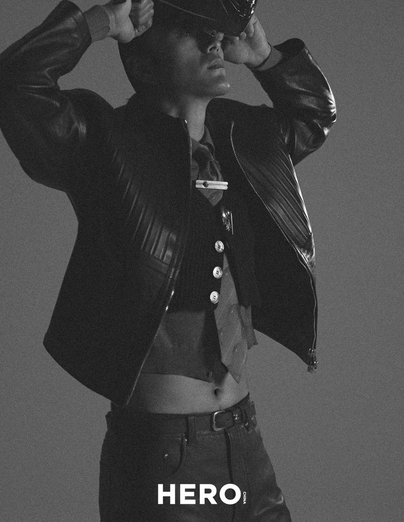 Model wearing a leather jacket and waistcoat adjusting a cap against a neutral grey studio backdrop.