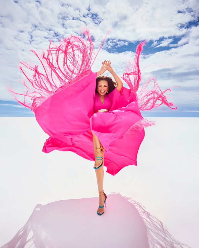 Thal5 in a flowing hot pink fringe dress leaping against a bright white ground under a cloudy blue sky.