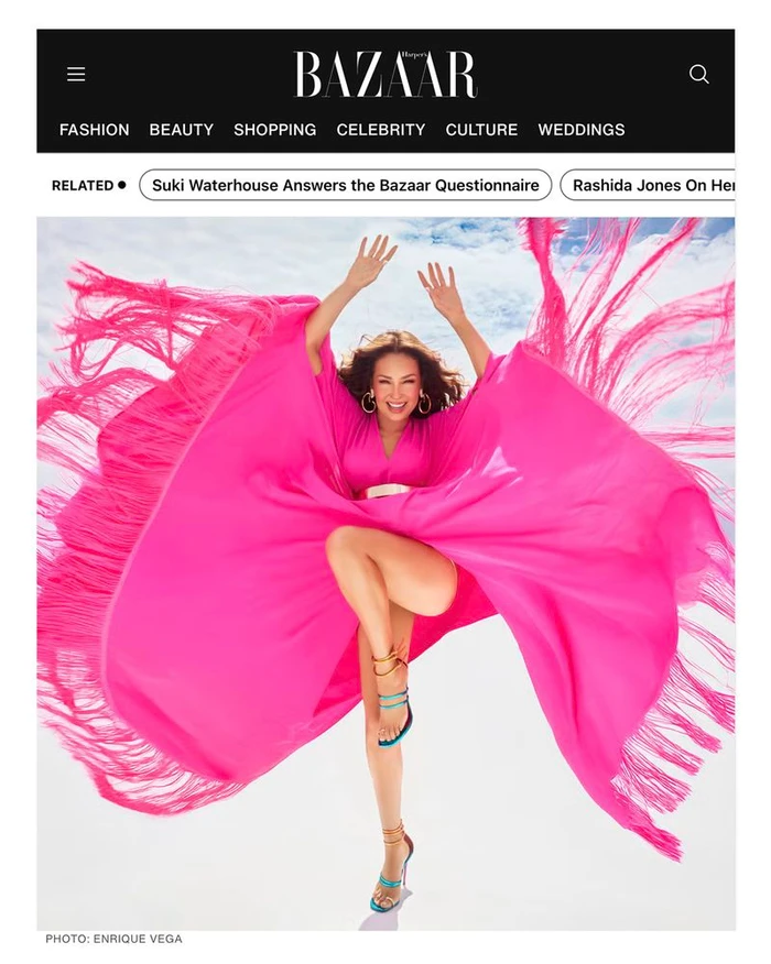 Thalía wearing a vibrant pink fringed dress leaping with arms raised against a bright sky background