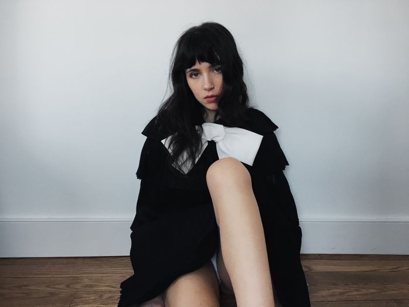 Clairo seated against a white wall wearing a black garment with a large white bow