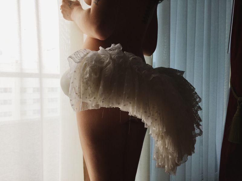 Partial view of a woman wearing a ruffled white lace skirt standing by a bright window with sheer curtains.