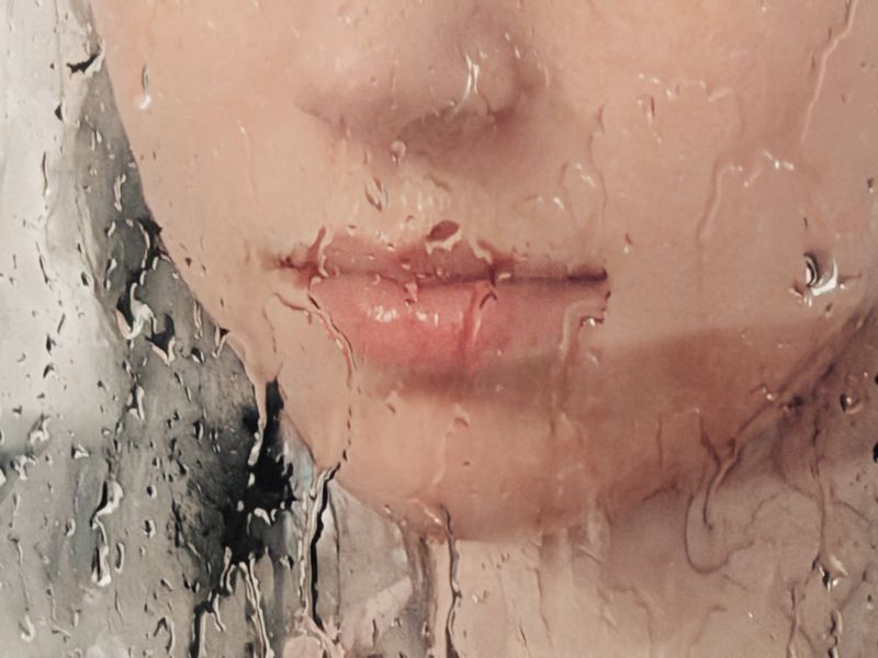 Close-up of a person's lower face behind a glass pane covered in water droplets and condensation.
