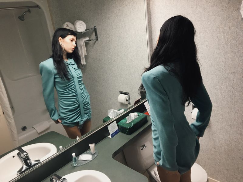Woman in a teal ruched minidress looking at her reflection in a hotel bathroom mirror for HommeGirls