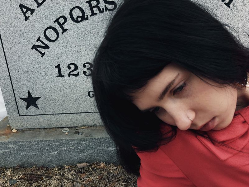 Close-up portrait of a woman with dark hair and a red top leaning against a large grey tombstone with printed letters and a star symbol.