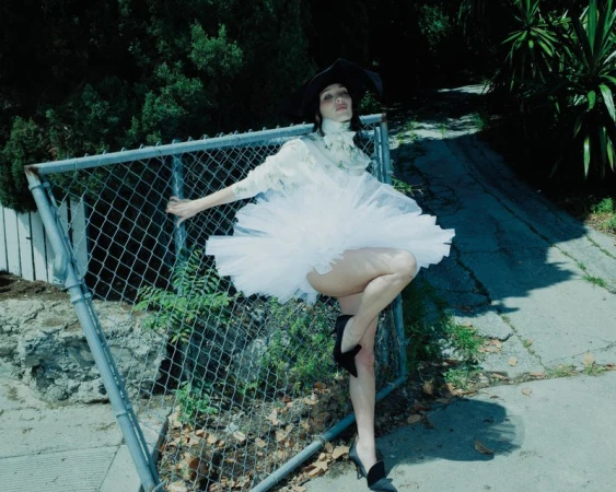 Maddie Ziegler wearing a white tulle dress and black hat leaning against a chain-link fence outdoors