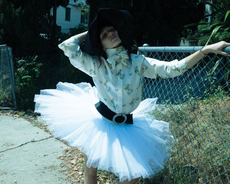 Maddie Ziegler wearing a floral blouse and white tulle tutu skirt posing by a chain-link fence for Rollacoaster Magazine.
