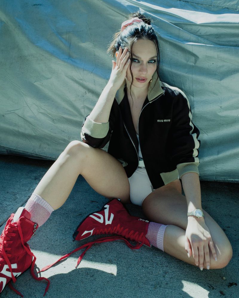 Maddie Ziegler sitting outdoors wearing a black track jacket, white bodysuit, pink socks, and red high-top boots.