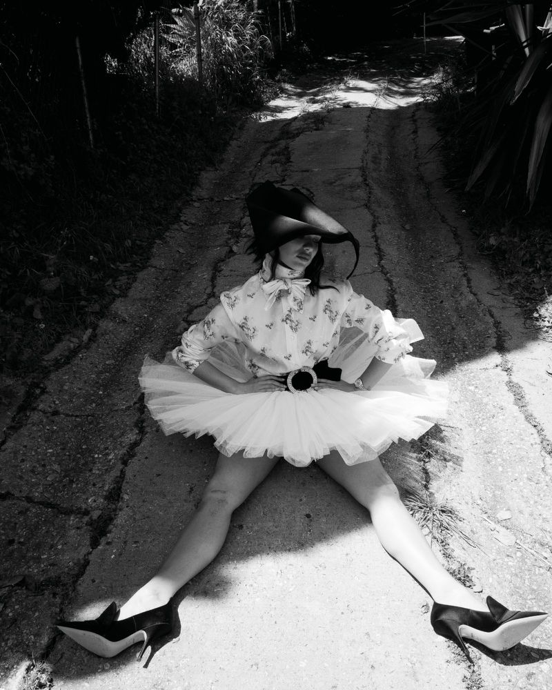 Maddie Ziegler in a white tutu and patterned blouse sitting on a rural asphalt path for Rollacoaster Magazine.