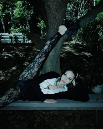 Maddie Ziegler lying on a wooden bench beneath a tree with one leg extended high, wearing black lace leggings and a black and white top.
