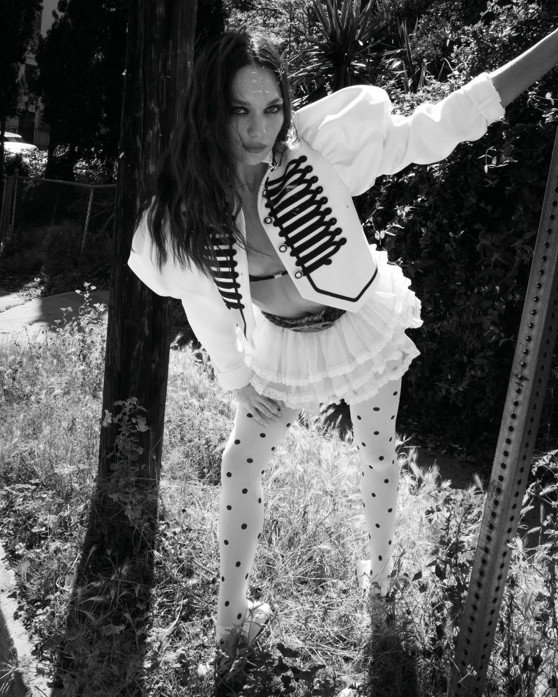Maddie Ziegler in a white military jacket and polka dot tights leaning outdoors for Rollacoaster Magazine