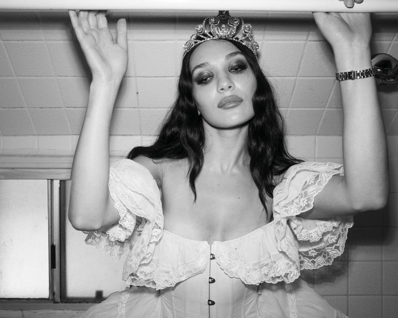 Maddie Ziegler wearing an ornate tiara and white off-the-shoulder lace dress in a tiled bathroom for Rollacoaster Magazine.