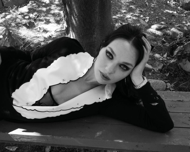 Maddie Ziegler lying on a wooden bench outdoors wearing a dark top with a white scalloped collar for Rollacoaster Magazine.
