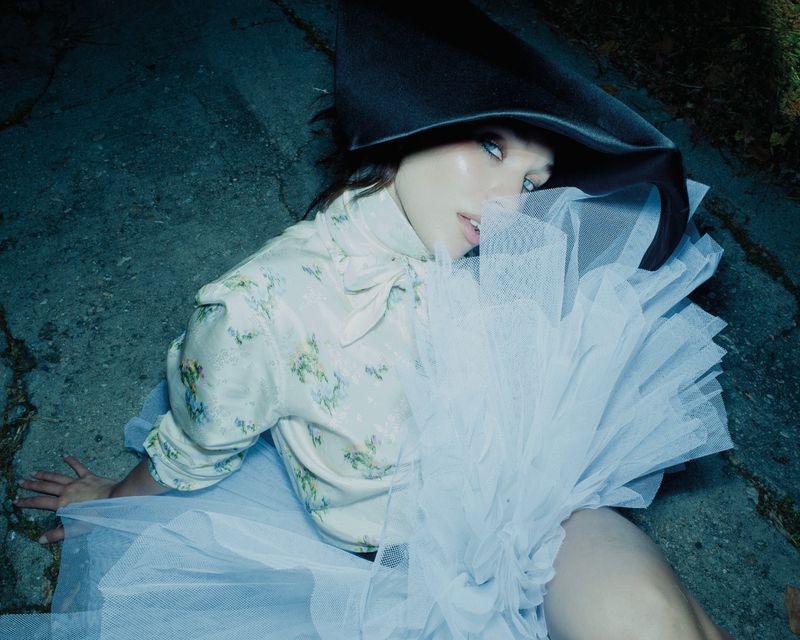 Maddie Ziegler wearing a large black hat, floral blouse, and white tulle skirt while seated outdoors on concrete.