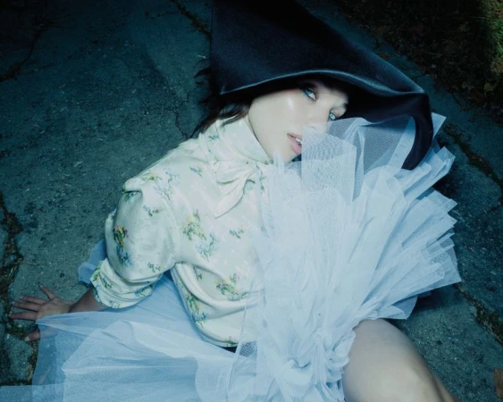 Maddie Ziegler wearing a large black hat, floral blouse, and white tulle skirt while seated outdoors on concrete.