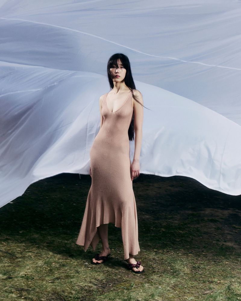 Model wearing a long beige slip dress standing outdoors with billowing white fabric in the background