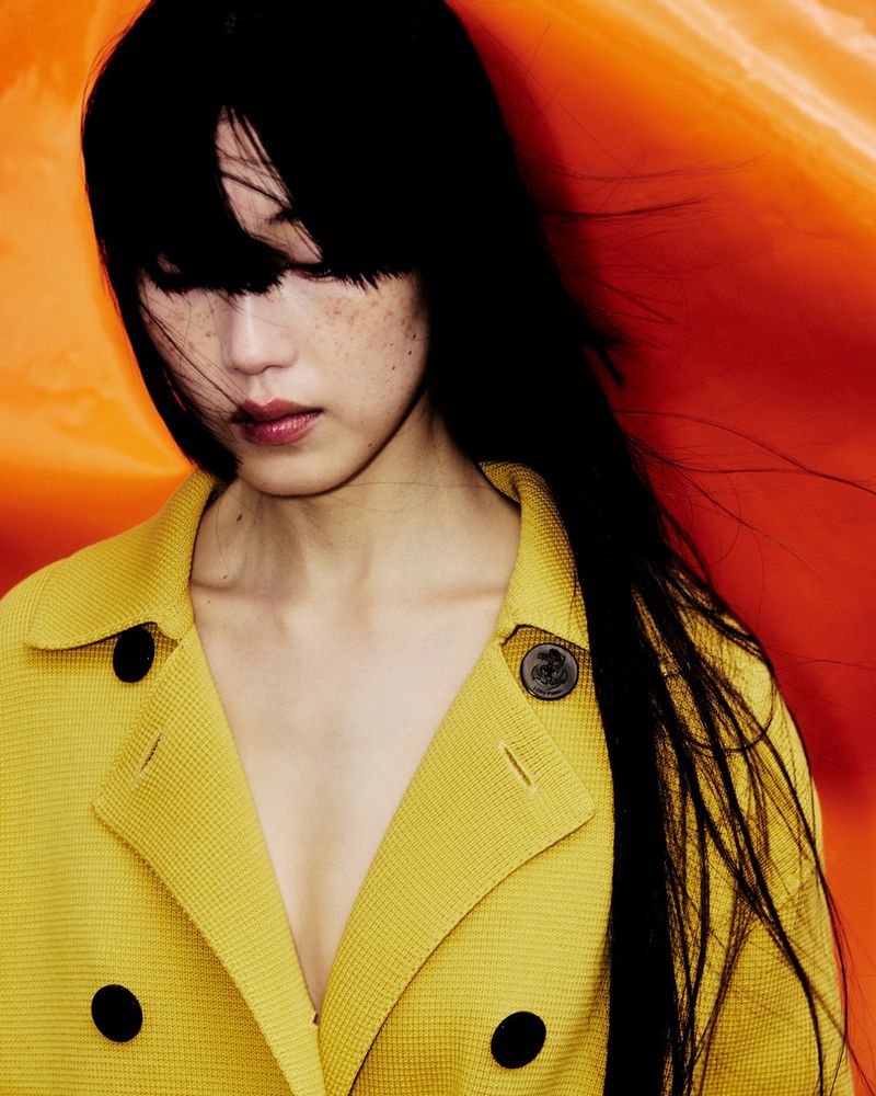 Model with long dark hair and freckles wearing a yellow textured coat against an orange backdrop for 10 Magazine Deutsch