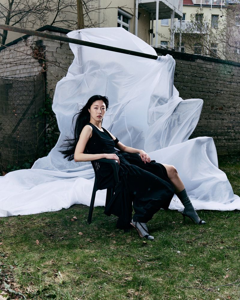 Model in black sleeveless dress seated on a black chair against a large draped white fabric backdrop in an outdoor garden.