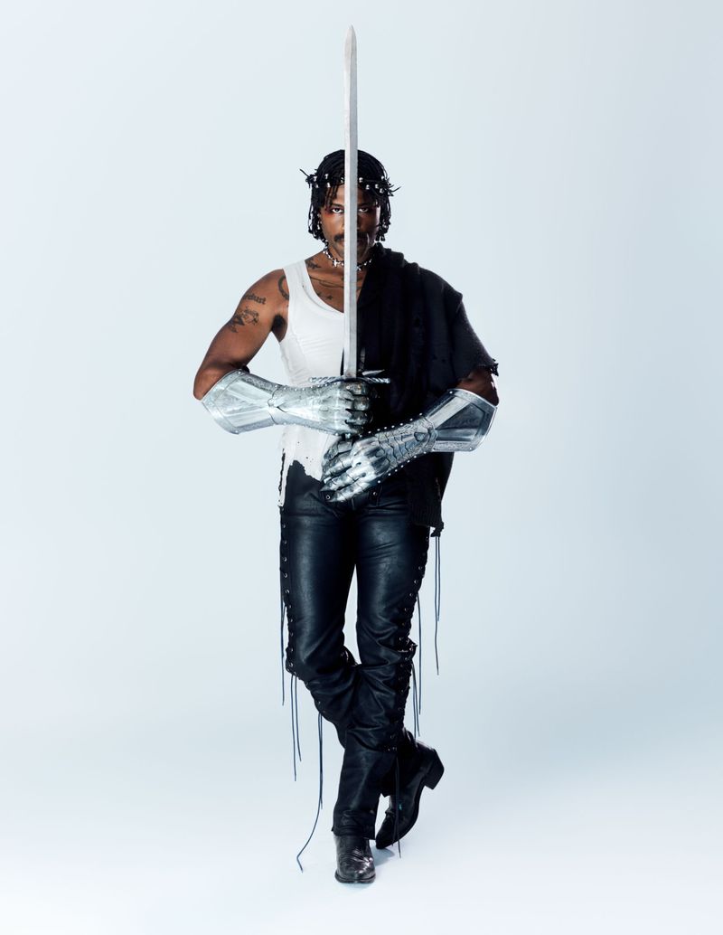 Man holding a sword vertically in front of his face, wearing silver gauntlets, a crown of thorns, a white tank top, and black leather pants