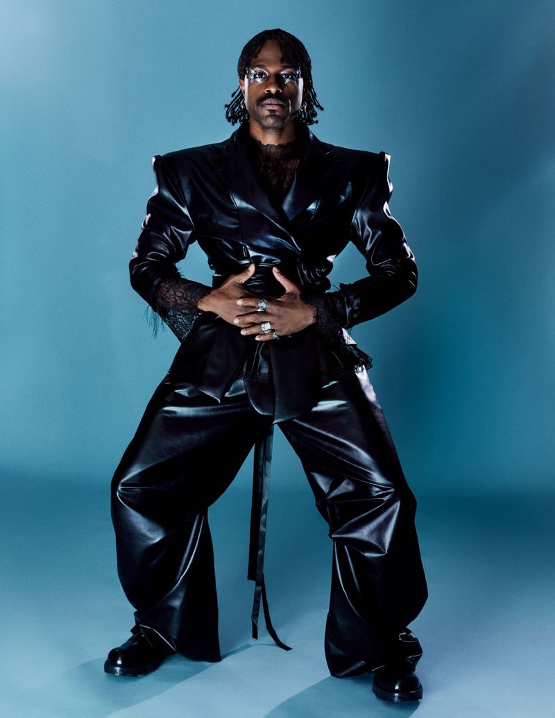 Adult male fashion model poses in a black pleather suit with dramatic shoulder pads on a light-blue gradient background in studio lighting.