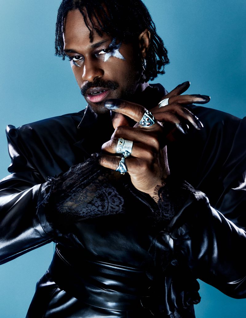 Portrait of Duckwrth wearing a black satin shirt with dramatic star eye makeup and silver statement rings against a blue background