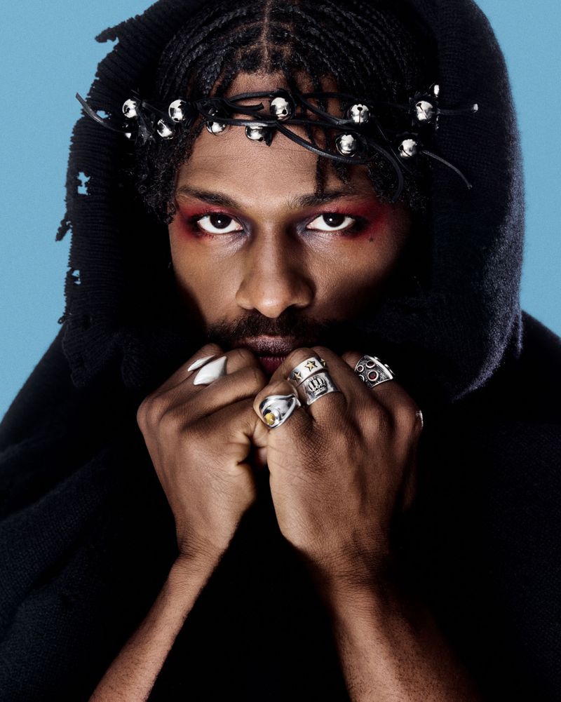 Close-up portrait of a Black man with braided hair wearing a dark hooded garment, silver rings, and red eye makeup against a blue backdrop