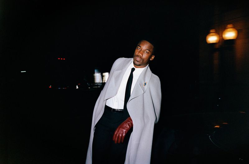 Giveon wearing a light grey coat, white shirt, black tie, and brown leather gloves against a dark nighttime backdrop.