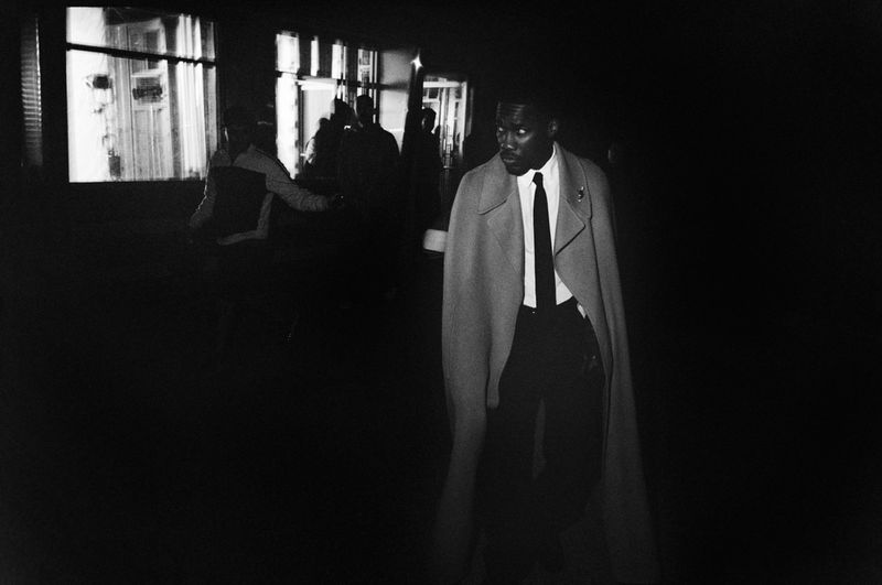 Monochrome portrait of a black man wearing a long coat and tie walking in a dark interior with illuminated windows
