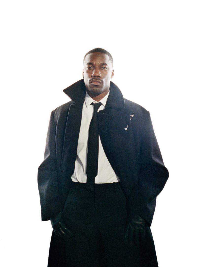 Black man wearing a dark coat, white shirt, black tie, and gloves against a white background