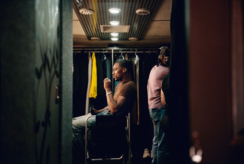 Black man seated in a dressing room looking sideways, with clothing racks and lit ceiling fixtures in the background.