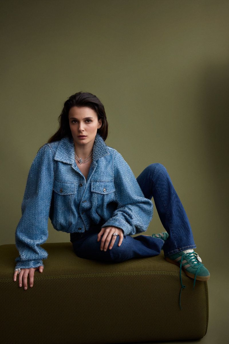 Woman in textured blue jacket and jeans sitting on olive green block against matching backdrop