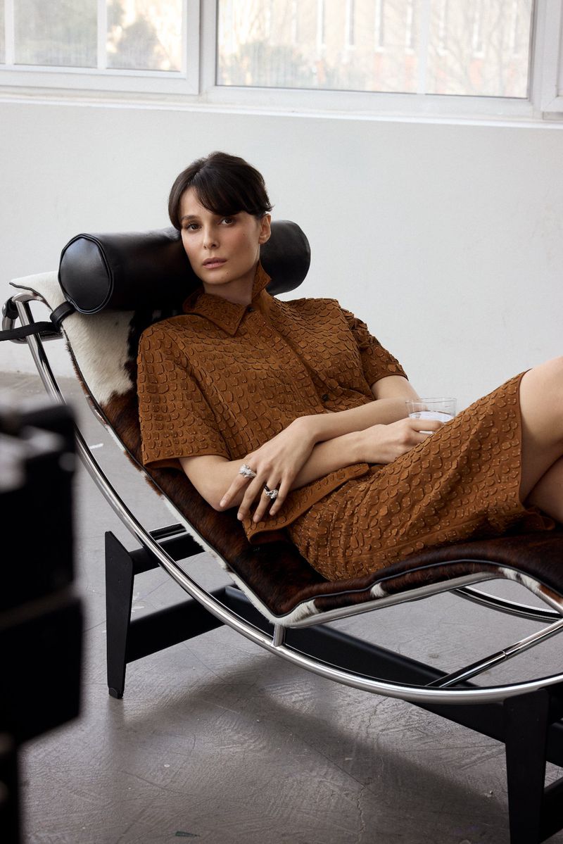 Woman in brown textured shirt and skirt reclining on a black leather lounge chair in an indoor setting.
