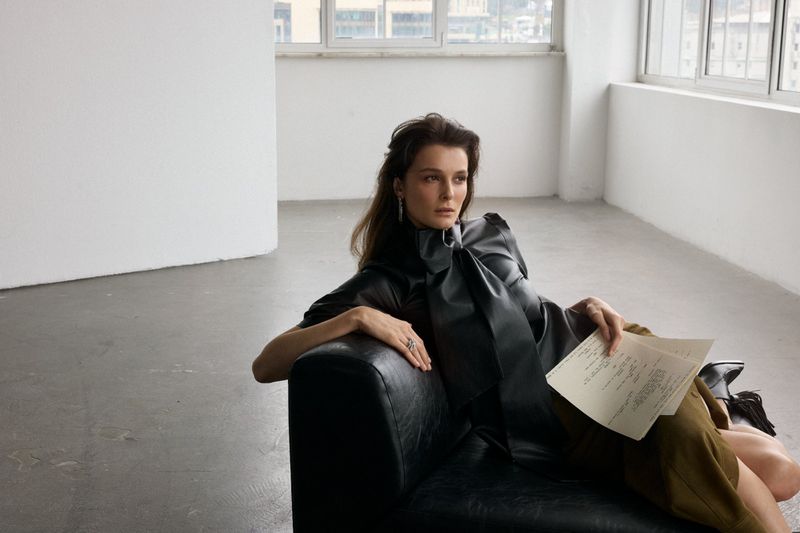 Woman in dark blouse and olive skirt holding papers while seated on a black leather armchair indoors