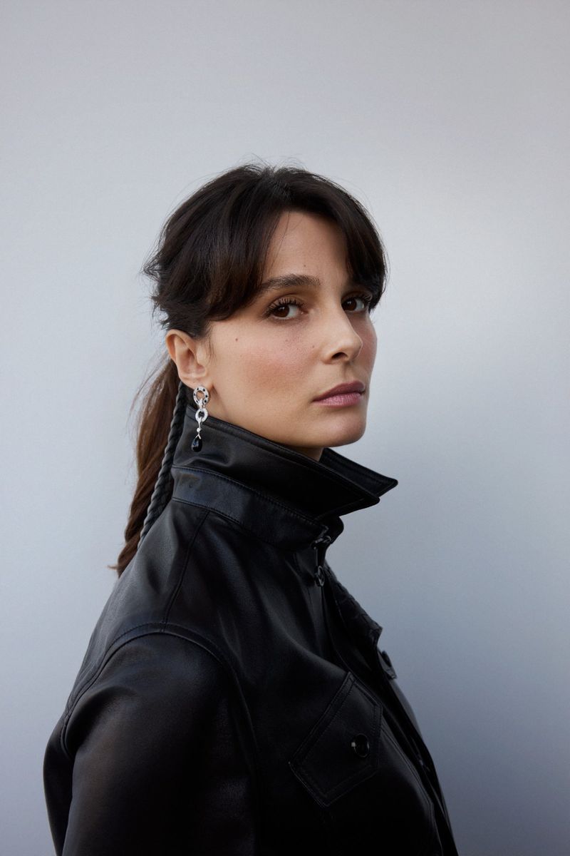 Woman in black jacket with braided hair and dangling earrings poses against a neutral background in a medium portrait framing.
