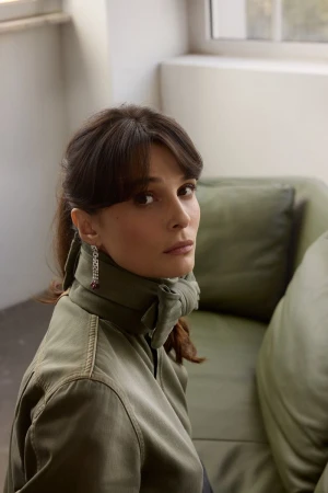 Busra Develi wearing an olive green collared jacket and drop earrings, seated near a green leather sofa.