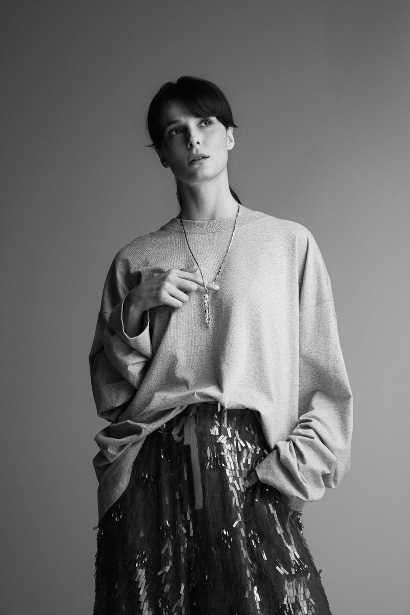 Black and white portrait of a woman wearing an oversized sweater and a pendant necklace against a plain grey backdrop.