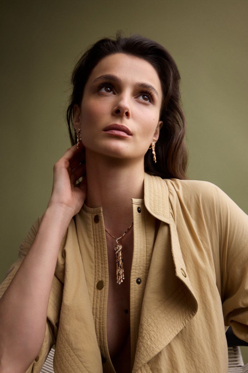 Portrait of actress Busra Develi wearing a tan shirt and gold tassel necklace against a solid olive background for Elle Turkiye.