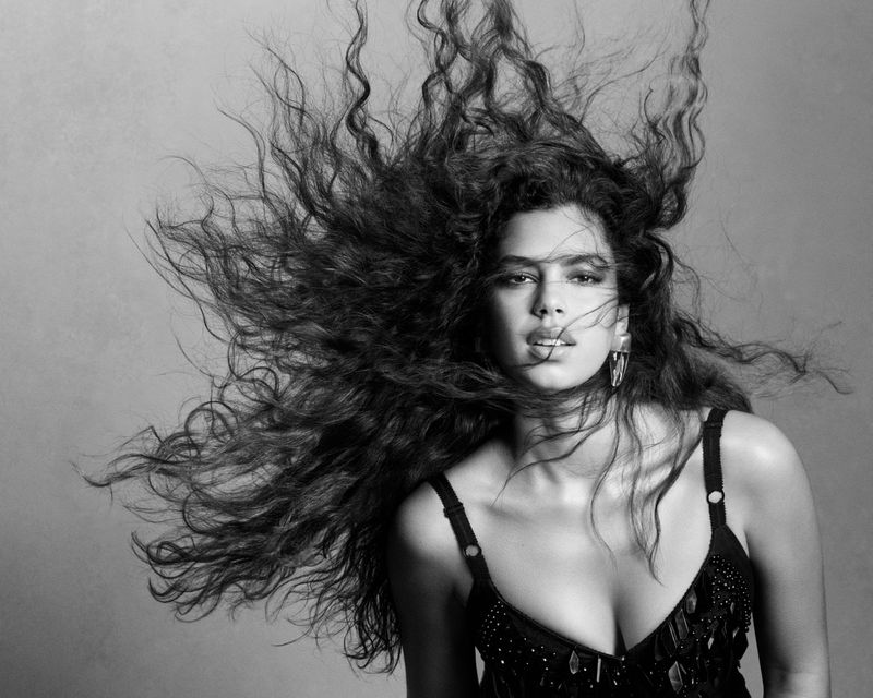 Black and white portrait of an adult woman with long, curly hair dynamically styled upwards. She wears a dark top with thin straps.