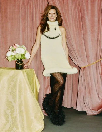 Brooke Shields wearing a cream knit sleeveless dress with a black smiley face motif and fluffy black shoes against pink drapes.