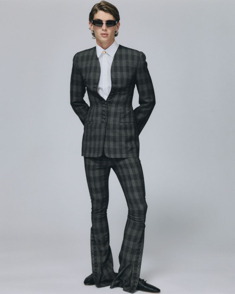 Model in dark plaid suit with flared trousers and sunglasses against a grey studio background for ODDA Magazine