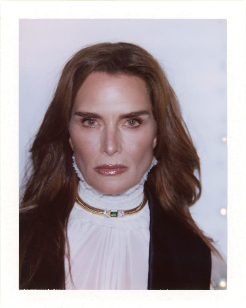 Portrait of Brooke Shields wearing a white ruffled blouse and gold choker necklace against a white backdrop.