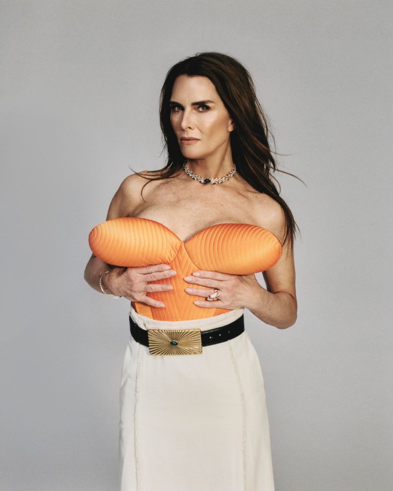 Brooke Shields wearing an orange structured strapless top and white skirt with a black belt against grey seamless background