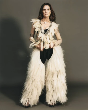 Brooke Shields wearing a feathered top and fringed chaps against a grey studio backdrop for ODDA Magazine