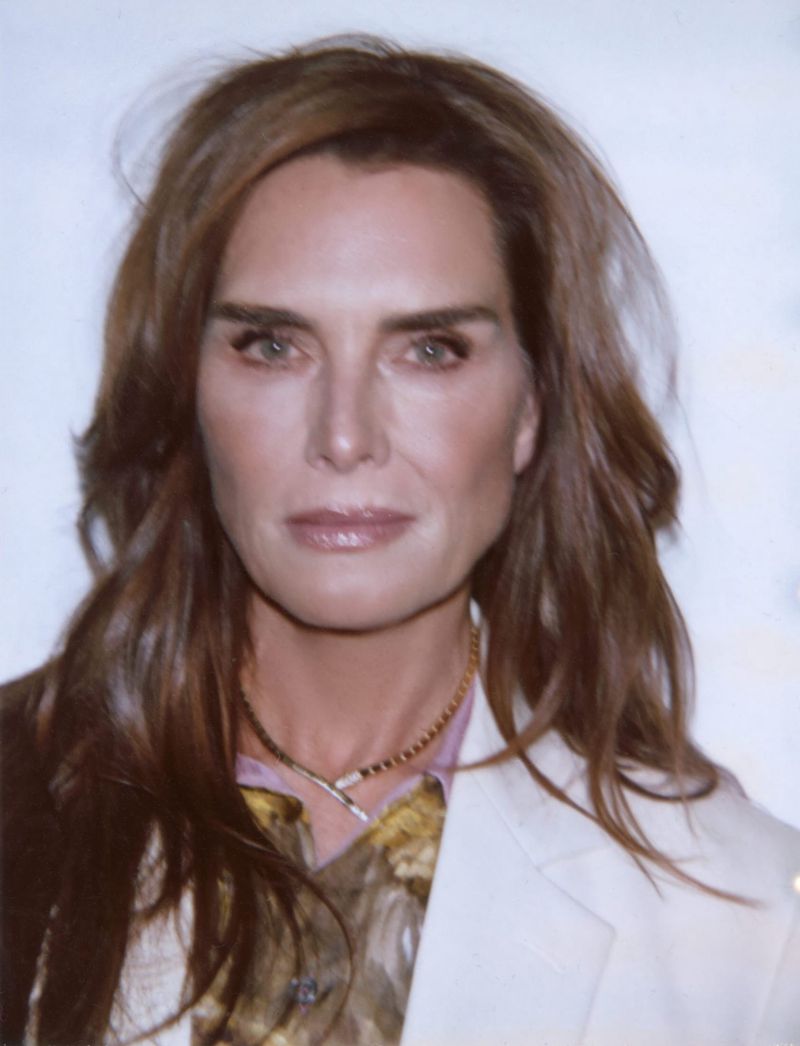 Portrait of Brooke Shields with long brown hair wearing a white jacket and gold necklace against a plain background.