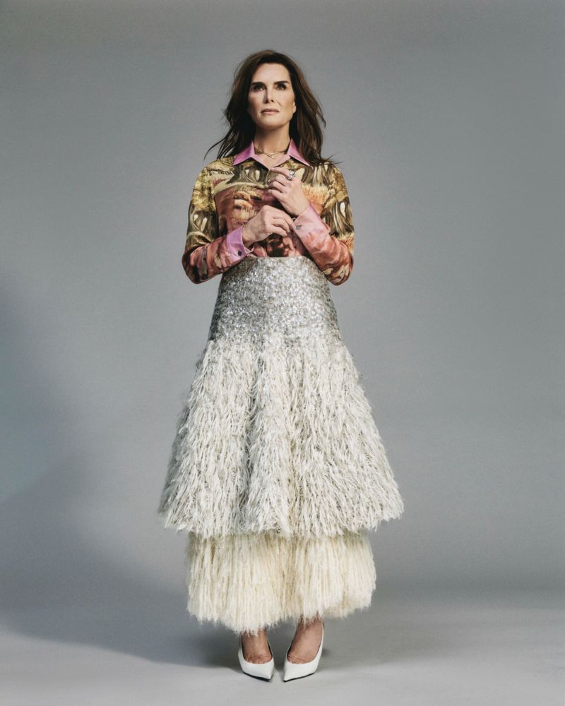 Studio shot of adult woman with dark hair wearing patterned top, sequined skirt, and heels against grey backdrop in full-length portrait.