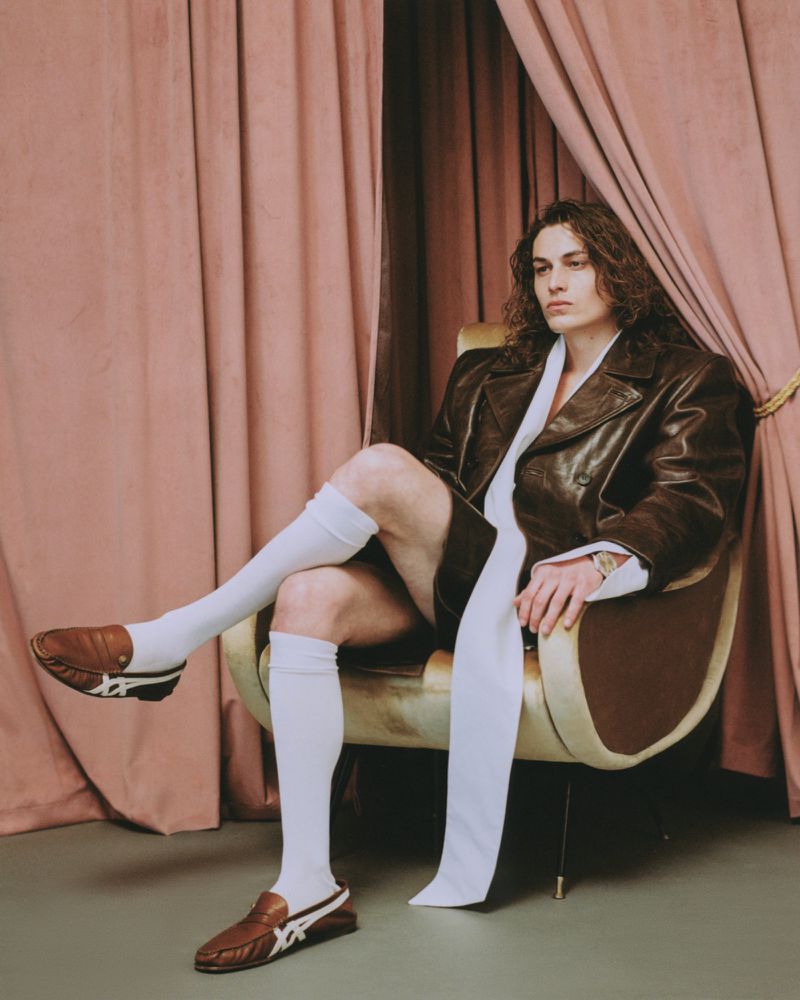 Model with long curly hair wearing a brown leather jacket and white knee-high socks seated in an armchair