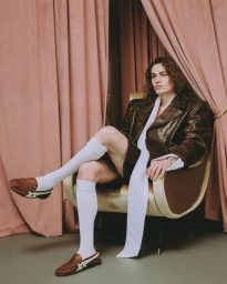 Model with long curly hair wearing a brown leather jacket and white knee-high socks seated in an armchair