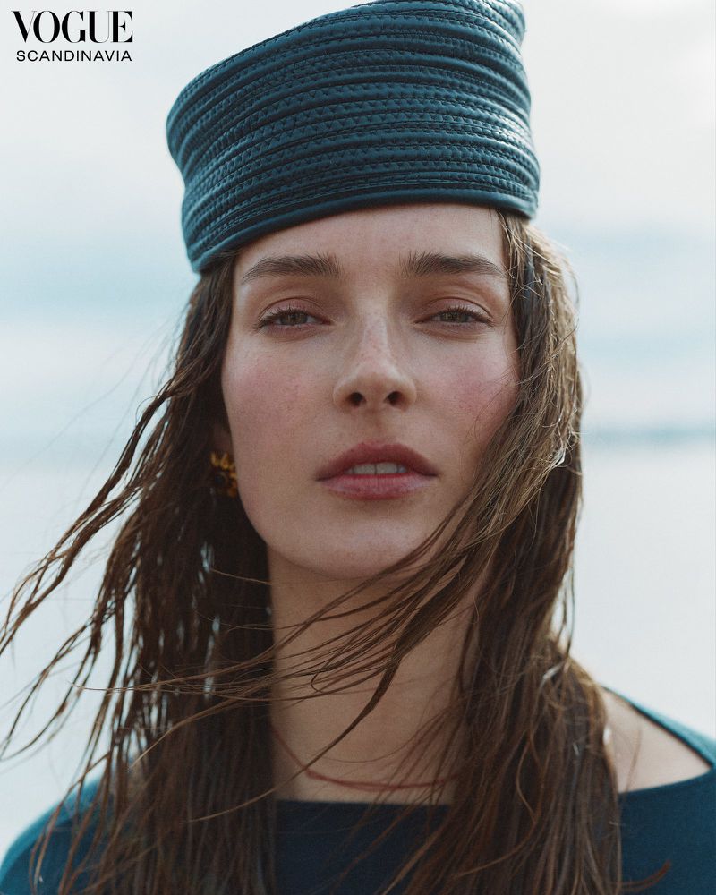 Close-up of a woman wearing a blue woven hat and matching top, posing in front of a blurred ocean background. Fashion editorial style.