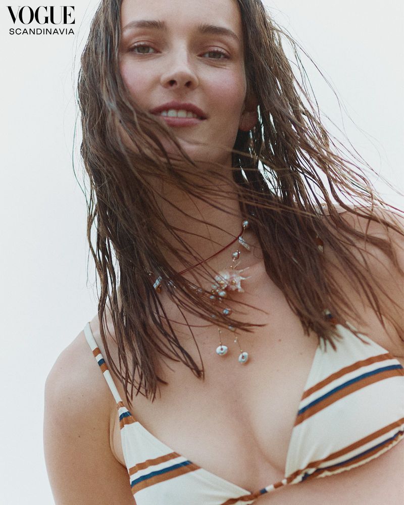 Close-up shot of a woman with wet brown hair and a striped bikini top, accessorized with a delicate pearl necklace against a light background.
