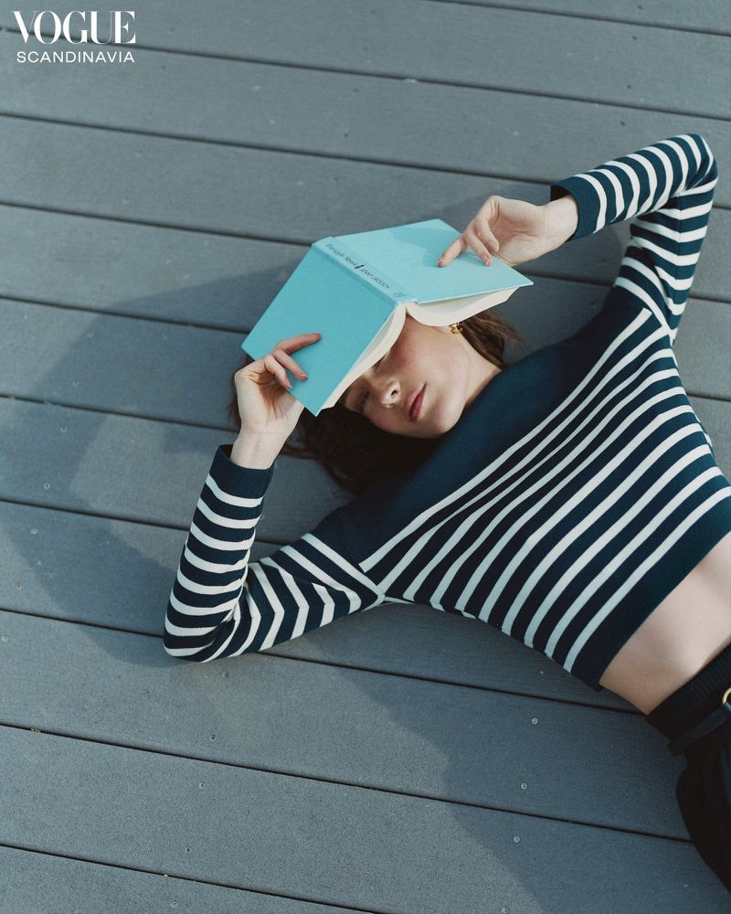 A woman with dark hair, wearing a striped top, lies on a grey wooden surface with a light blue book covering her face.