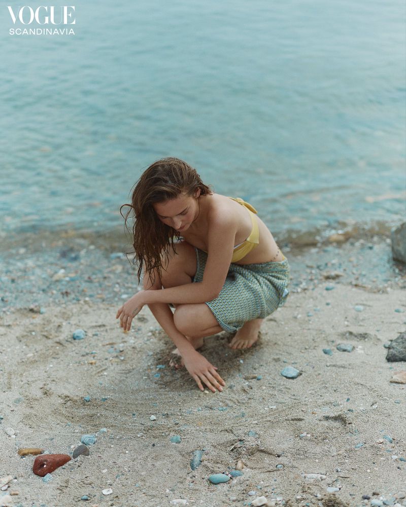 A young woman wearing a yellow bikini top and textured knit shorts squats on a sandy beach next to the ocean on a cloudy day.